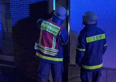 Zwei Einsatzkräfte in THW-Uniformen untersuchen nachts die Außenfassade eines mit Blaulicht beleuchteten Gebäudes nach einem möglichen Gasaustritt oder einer Explosion.