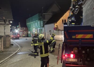 Feuerwehrleute in Schutzkleidung reagieren nachts auf einen möglichen Gasaustritt in der Nähe eines Lastwagens in einer Wohnstraße. Eine Person befindet sich auf der Ladefläche des Lastwagens, andere koordinieren die Maßnahmen nach Berichten über eine Explosion.