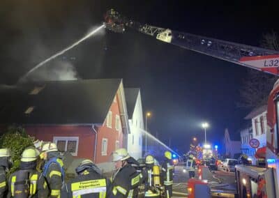 Feuerwehrleute löschen nachts mit Schläuchen und Drehleiter einen Dachstuhlbrand an einem Wohnhaus; vom Dachstuhl ist Rauch zu sehen.