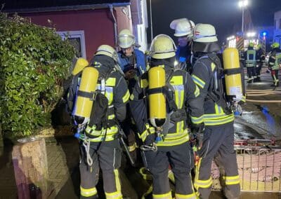 Eine Gruppe Feuerwehrleute in voller Montur mit Sauerstoffflaschen steht nachts auf einer nassen Straße in der Nähe eines Hauses, beleuchtet von Notlichtern nach einem Dachstuhlbrand.