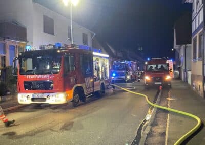 Feuerwehrautos und Einsatzfahrzeuge parken nachts in einer Wohnstraße. Auf der nassen Straße sind während eines Einsatzes nach einem Dachstuhlbrand Schläuche ausgelegt.