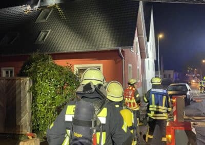 Feuerwehrleute in Schutzkleidung reagieren nachts auf einen Dachstuhlbrand. Auf dem Dach ist Rauch zu sehen, und über ihnen ist eine Drehleiter ausgefahren.