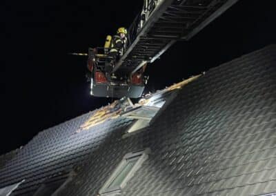 Ein Feuerwehrmann untersucht auf einer Drehleiter nachts den Dachschaden eines dunklen Hauses, der durch einen Dachstuhlbrand verursacht wurde. Um den beschädigten Abschnitt herum liegen Trümmer verstreut.