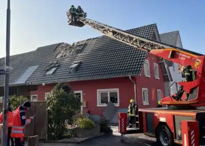 Feuerwehrleute nutzen eine Drehleiter, um den Dachschaden durch einen Dachstuhlbrand an einem roten Haus zu untersuchen, während Rettungskräfte in der Nähe auf der Straße stehen.