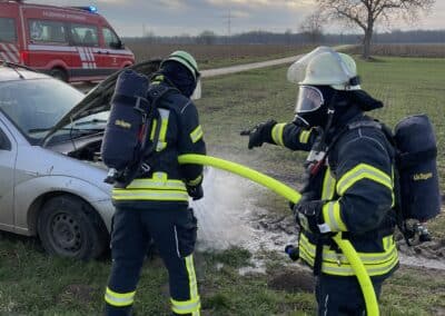 Zwei Feuerwehrmänner in Schutzanzügen löschen einen Brand unter der Motorhaube eines freistehenden PKW auf einer Wiese, während Rauchentwicklung aufsteigt und ein Feuerwehrauto in der Nähe geparkt steht.
