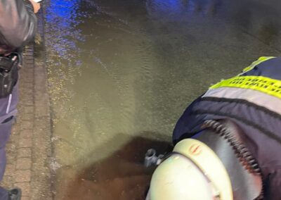 Ein Feuerwehrmann mit Helm arbeitet nachts in der Nähe einer überfluteten Straße nach einem Wasserrohrbruch, während eine andere Person in der Nähe steht und einen Notizblock hält.