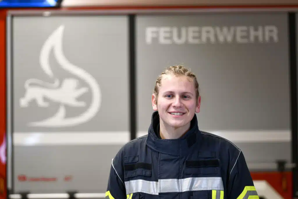 Ein Feuerwehrmann in Uniform steht lächelnd vor einem Feuerwehrauto, im Hintergrund sind das Wort „Feuerwehr“ und ein Flammensymbol zu sehen.