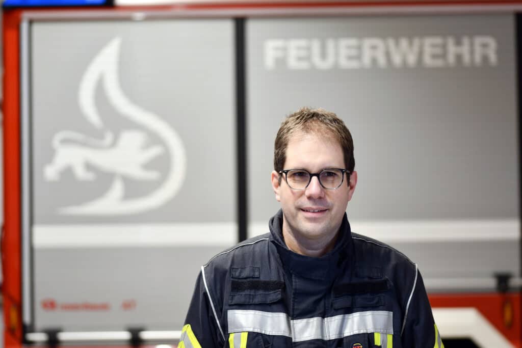 Ein Feuerwehrmann in Uniform steht in einem Gebäude vor einem Feuerwehrauto. Im Hintergrund sind das Wort FEUERWEHR und ein Feuersymbol zu sehen.