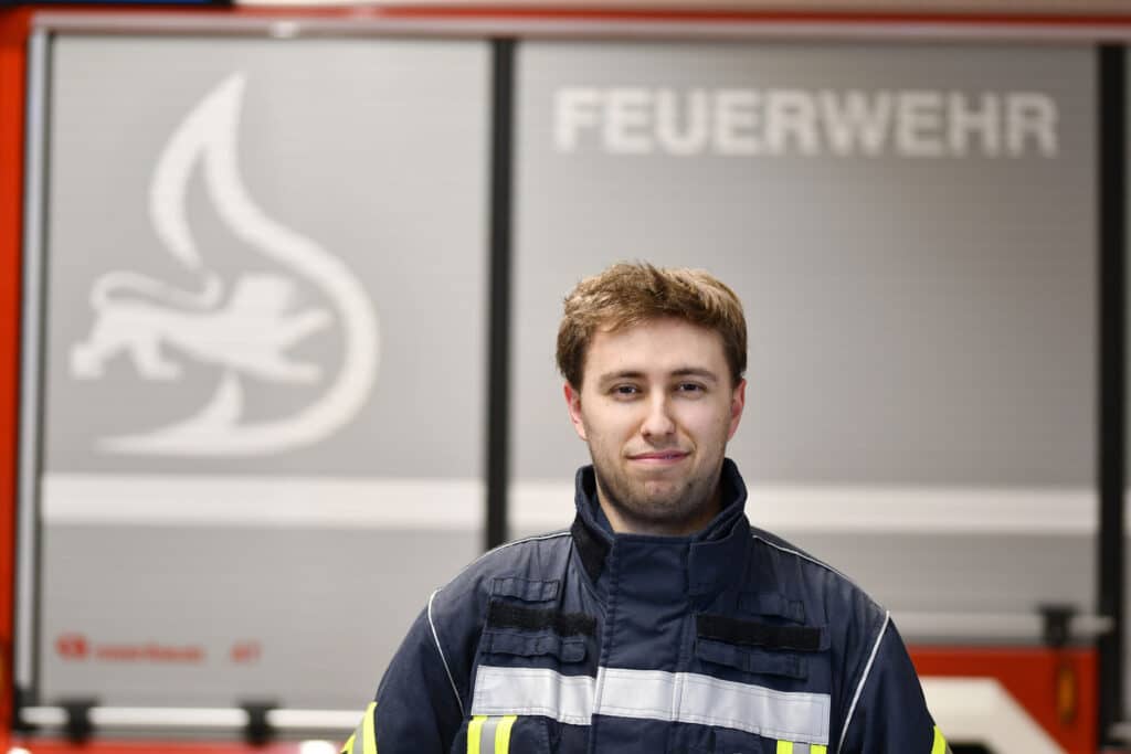 Ein Feuerwehrmann in Uniform steht vor einem Feuerwehrauto, im Hintergrund sind das Wort „Feuerwehr“ und ein Flammensymbol zu sehen.