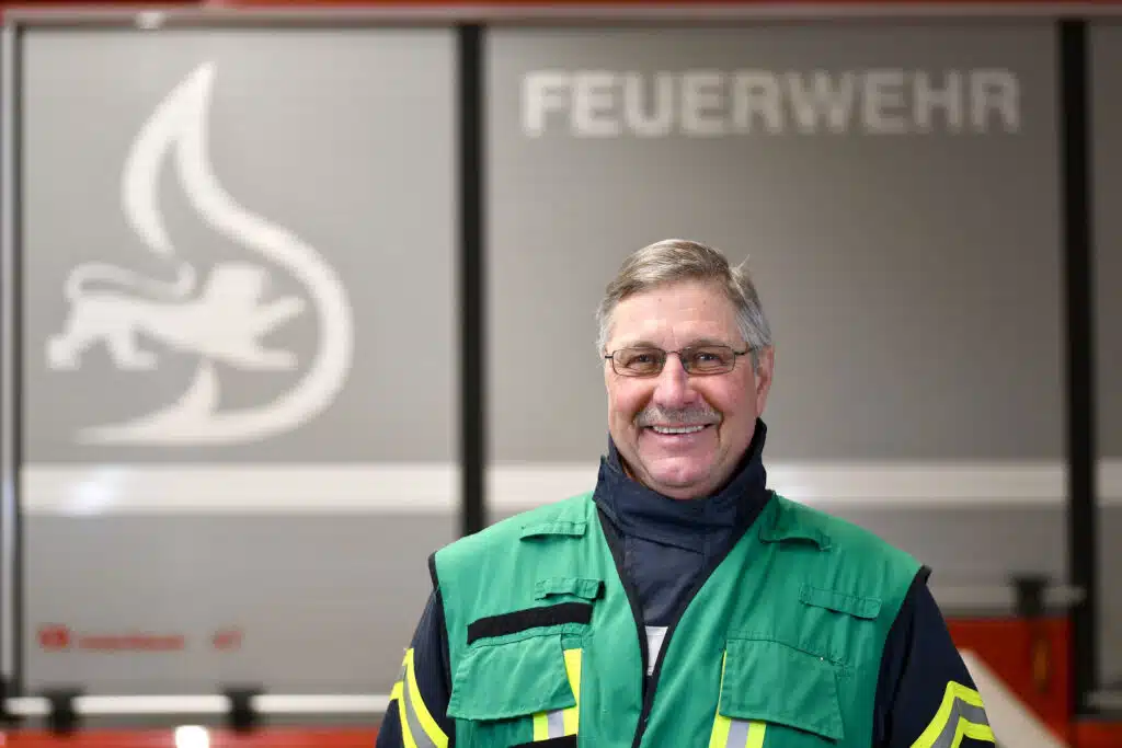 Lächelnder Mann in grüner Warnweste steht vor einem Feuerwehrauto mit der Aufschrift FEUERWEHR.