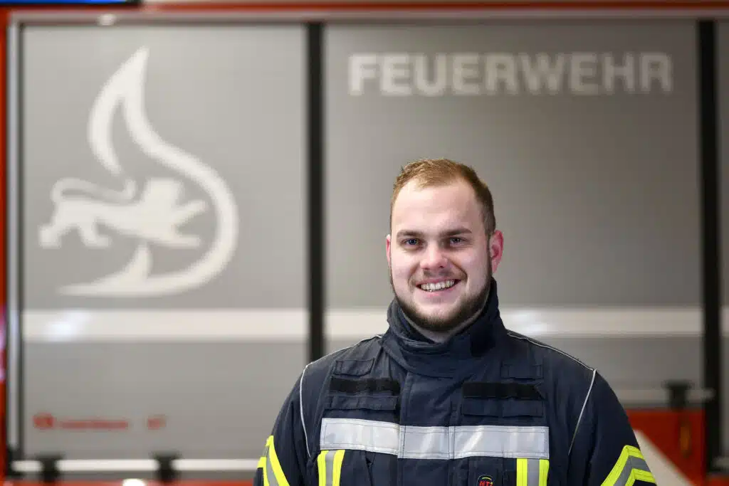 Ein Mann in Feuerwehruniform steht lächelnd vor einer Wand mit dem Wort FEUERWEHR und einem Feuersymbol.