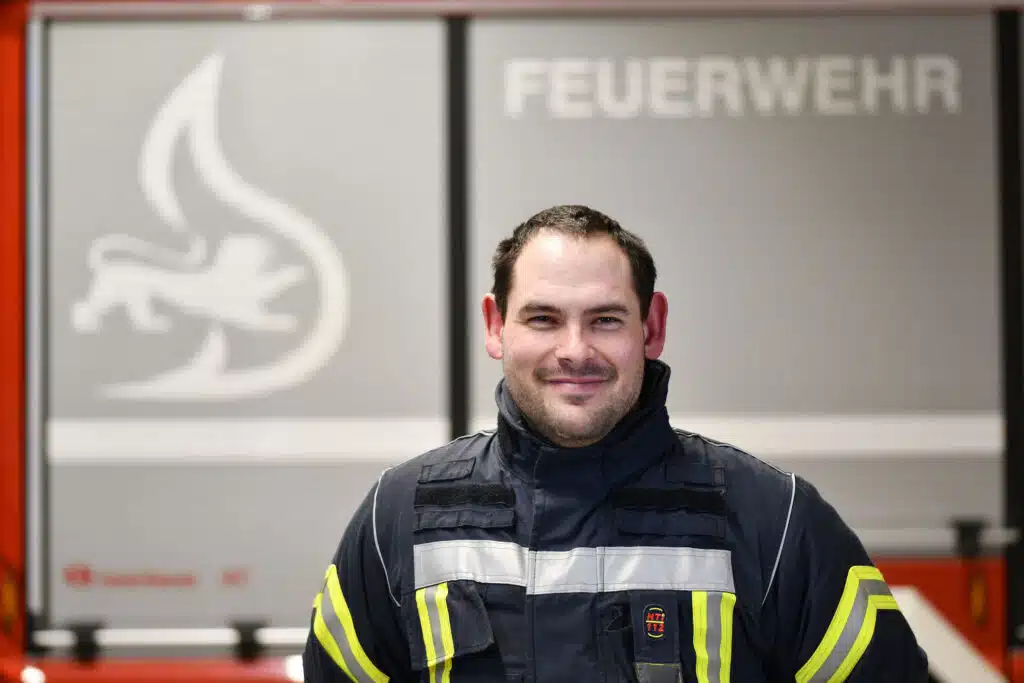 Ein Feuerwehrmann in Uniform steht vor einem Feuerwehrauto, im Hintergrund ist das Wort „Feuerwehr“ zu sehen.