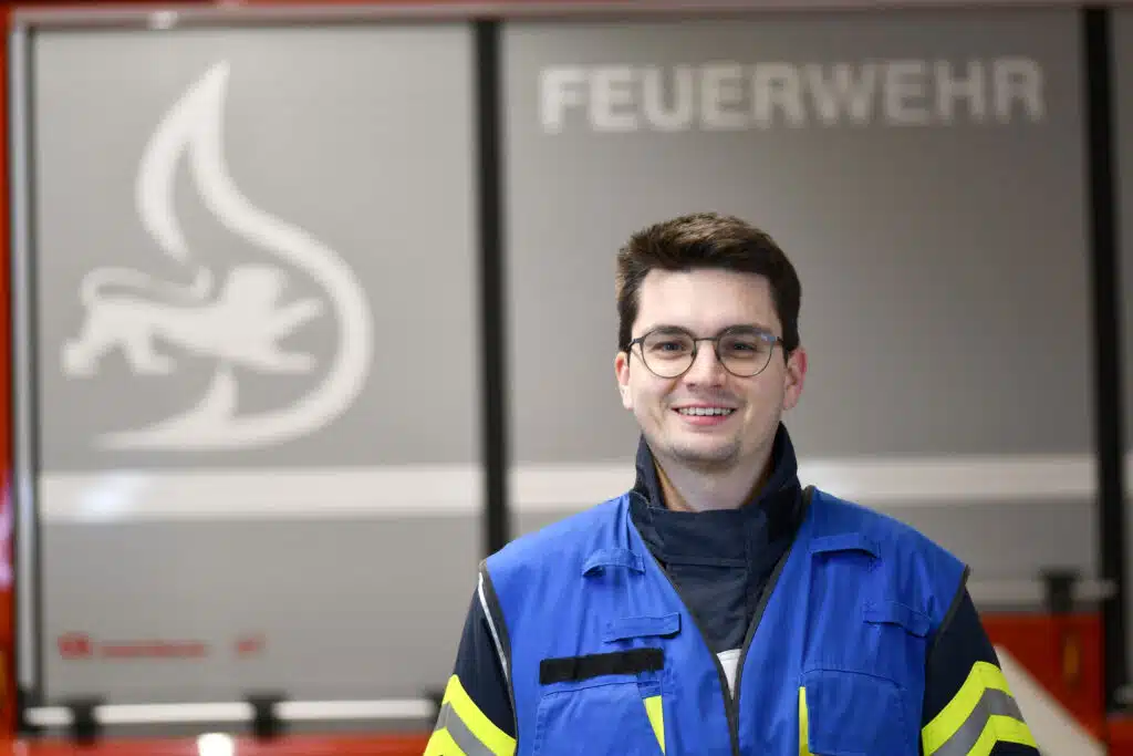 Ein Mann in blau-gelber Sicherheitsuniform steht vor einem Feuerwehrschild mit der Aufschrift FEUERWEHR.