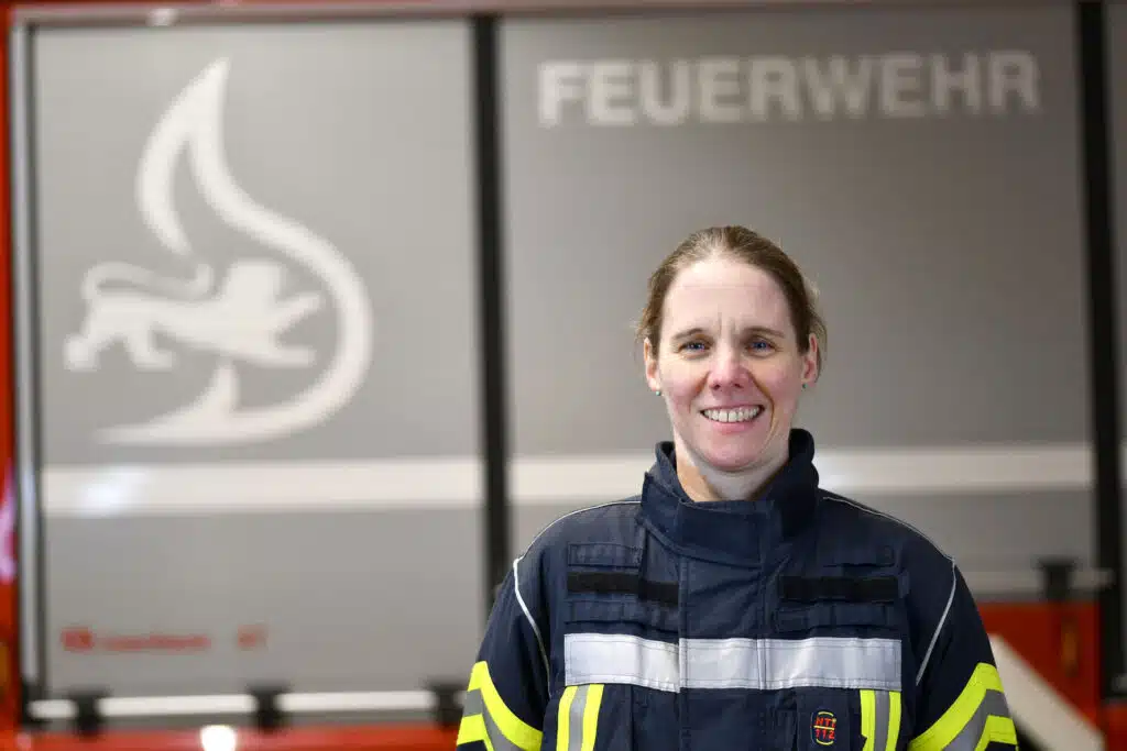 Eine Frau in Feuerwehruniform steht lächelnd vor einem Feuerwehrauto, im Hintergrund sind das Wort FEUERWEHR und ein Feuersymbol zu sehen.