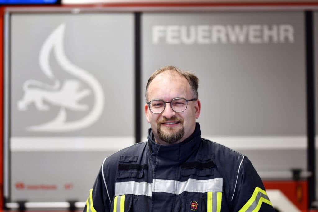 Ein Mann in Feuerwehruniform steht vor einem Feuerwehrauto, im Hintergrund ist das Wort „Feuerwehr“ zu sehen.