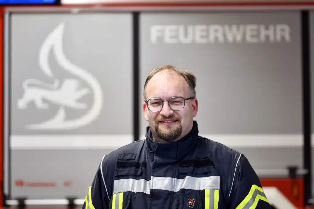 Ein Mann in Feuerwehruniform steht vor einem Feuerwehrauto, im Hintergrund ist das Wort „Feuerwehr“ zu sehen.