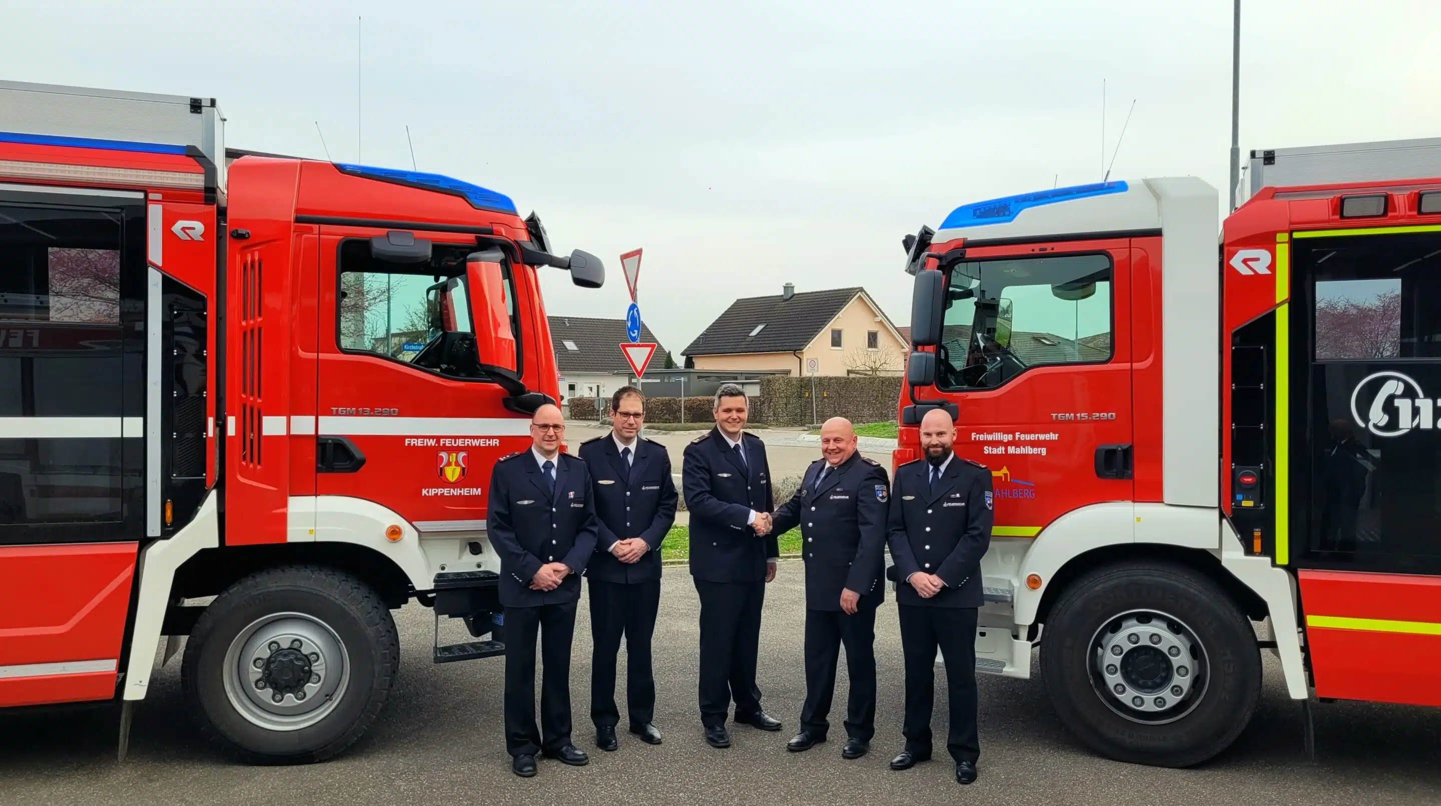 Fünf uniformierte Feuerwehrleute stehen zwischen zwei roten Feuerwehrautos, im Hintergrund sind Häuser und ein Verkehrsschild zu sehen.