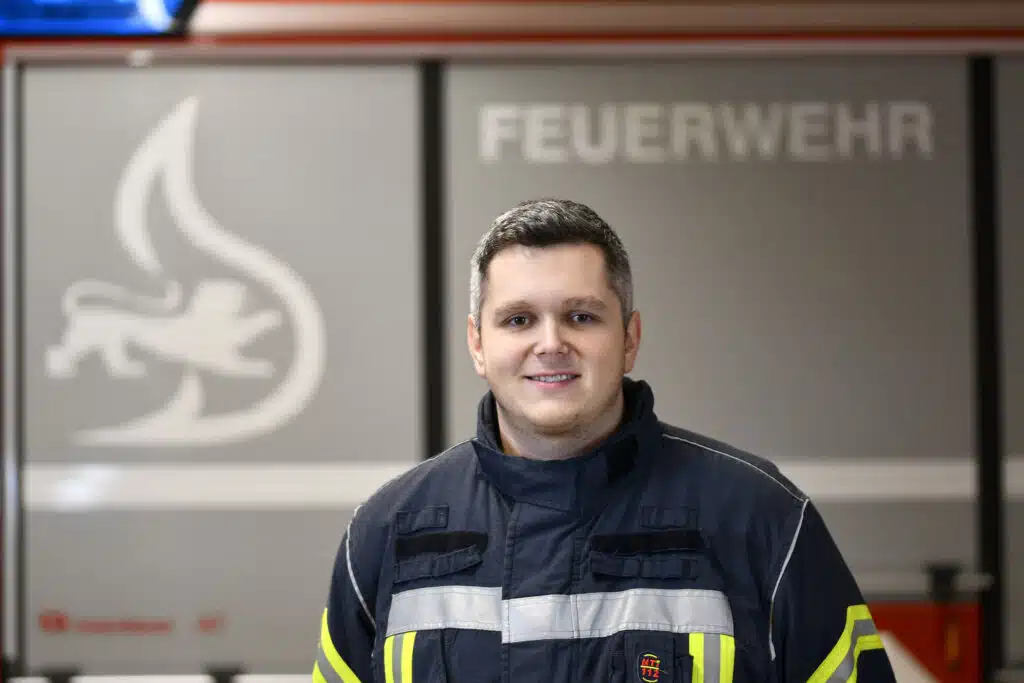 Ein Feuerwehrmann in Uniform steht vor einem Feuerwehrauto, im Hintergrund sind das Wort FEUERWEHR und ein Flammenlogo zu sehen.