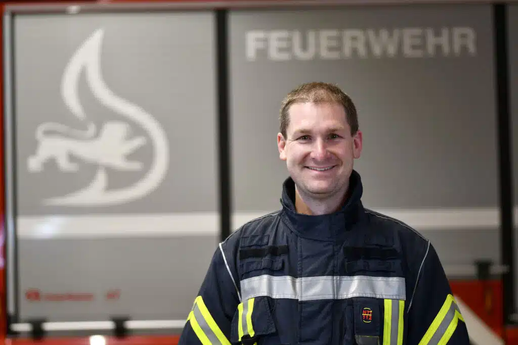 Ein Feuerwehrmann in Uniform steht lächelnd vor einem Feuerwehrauto, im Hintergrund ist das Wort FEUERWEHR zu sehen.