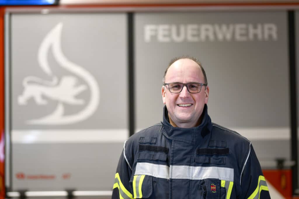 Ein Mann in Feuerwehruniform steht lächelnd vor einem Feuerwehrauto mit der Aufschrift „Feuerwehr“ und einem Flammensymbol im Hintergrund.