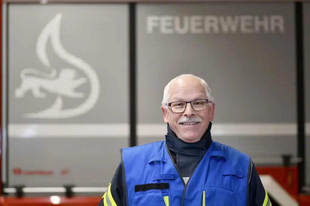 Ein älterer Mann mit grauem Haar und Brille, der eine blaue Sicherheitsweste trägt, steht im Innenbereich vor einer Wand mit der Aufschrift „FEUERWEHR“ und einem Flammen- und Löwensymbol.