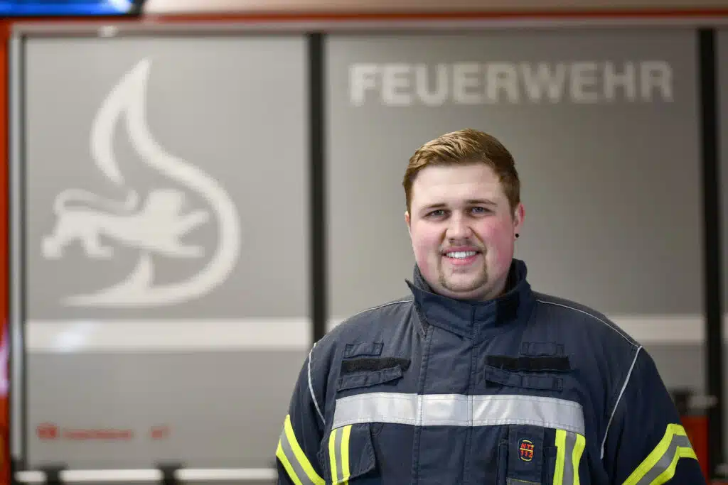 Ein Feuerwehrmann in dunkler Uniform mit Reflektorstreifen steht vor einer Wand mit der Aufschrift „Feuerwehr“ und einem Feuerwehrlogo.
