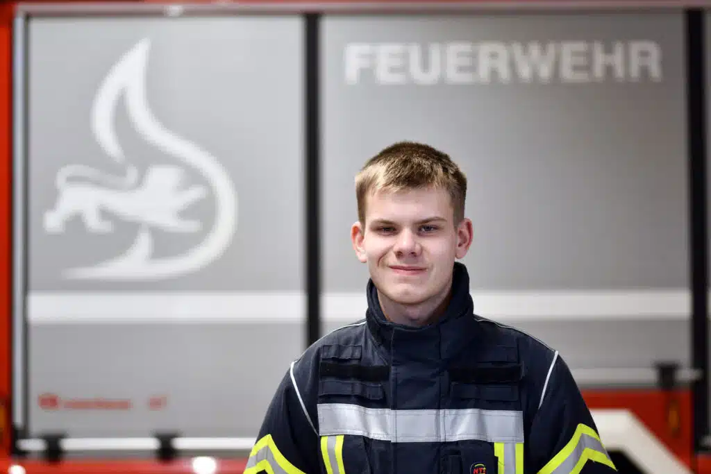 Ein junger Feuerwehrmann in Uniform steht vor der Tür einer Feuerwache, im Hintergrund sind das Wort FEUERWEHR und ein Feuersymbol zu sehen.