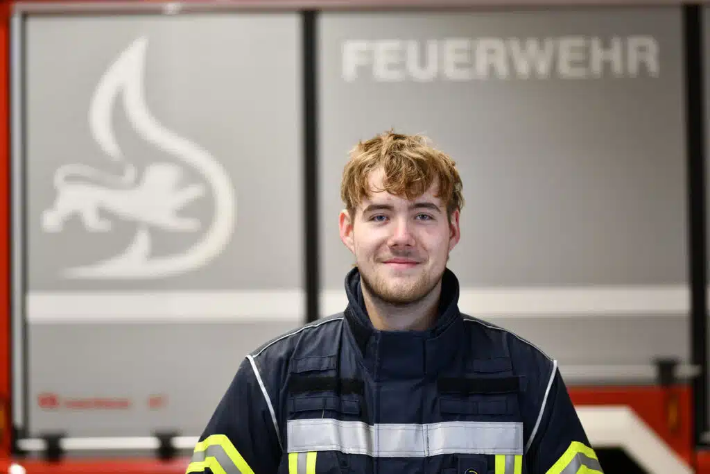Ein Feuerwehrmann in Uniform steht im Innenbereich vor einer Wand mit dem Wort FEUERWEHR und einem Flammensymbol.