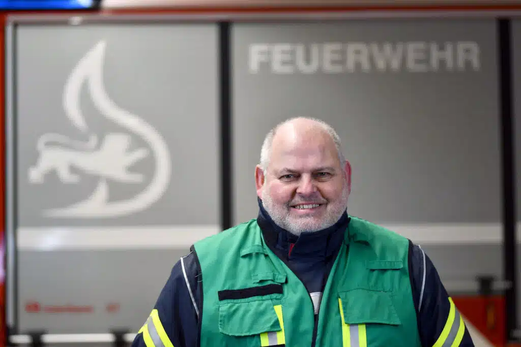 Ein Mann mit einer grünen Warnweste steht vor einer Wand mit der Aufschrift „Feuerwehr“ und einem Feuerwehrsymbol.