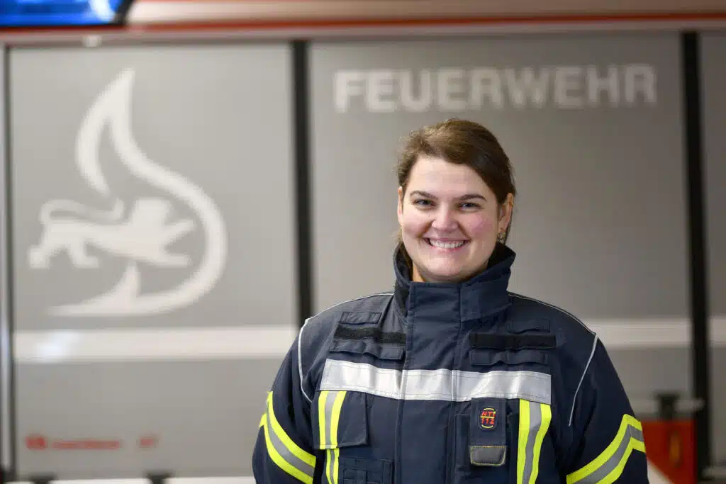 Eine Frau in Feuerwehruniform steht vor einem Feuerwehrauto mit der Aufschrift „Feuerwehr“.