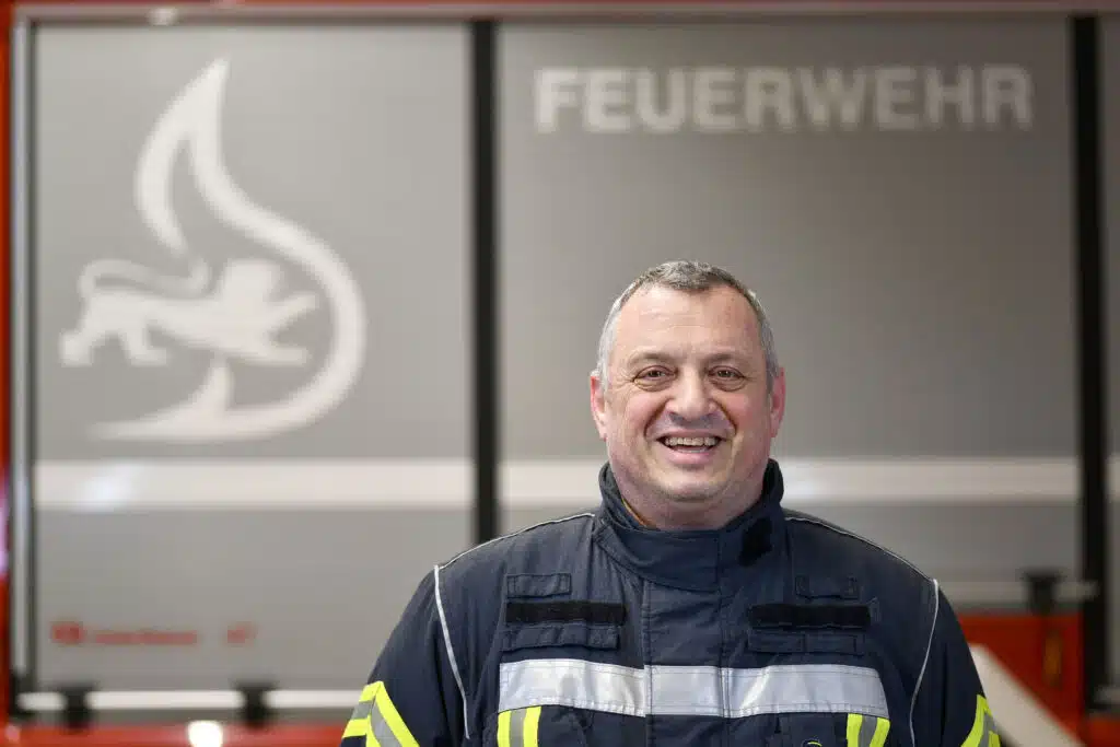 Ein Feuerwehrmann in Uniform steht lächelnd vor einem Feuerwehrauto mit der Aufschrift FEUERWEHR.