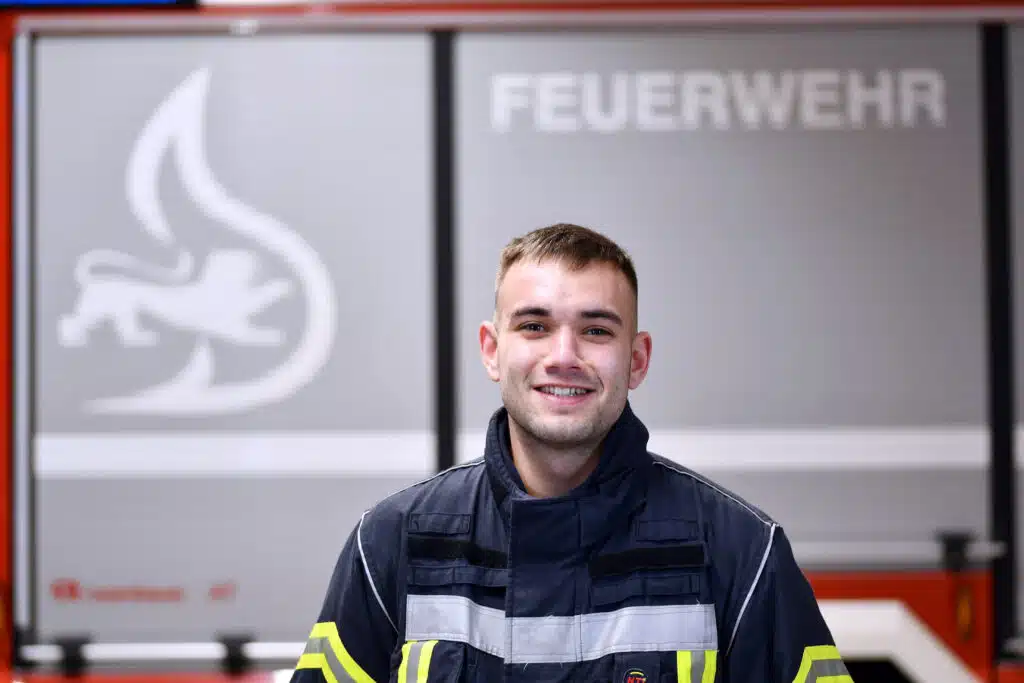Ein Feuerwehrmann in Uniform steht lächelnd vor einem Feuerwehrauto mit der Aufschrift „Feuerwehr“.